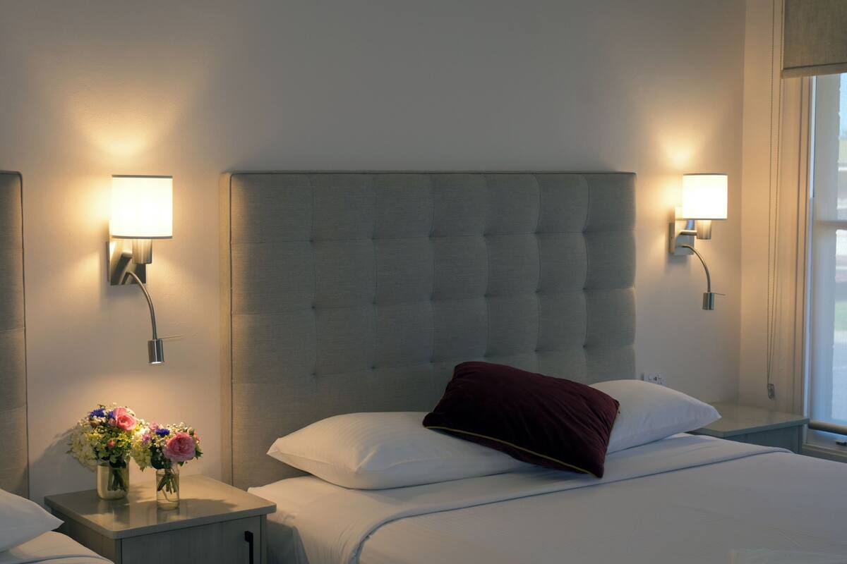 Habitació d'hotel amb un panell de capçal tapissat, dues làmpades de nit, una tauleta de nit amb un ram de flors i una finestra amb cortines.