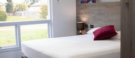 A bedroom with a large window, a bed with white bedding and a red pillow, a wall-mounted light, and a colorful abstract painting.