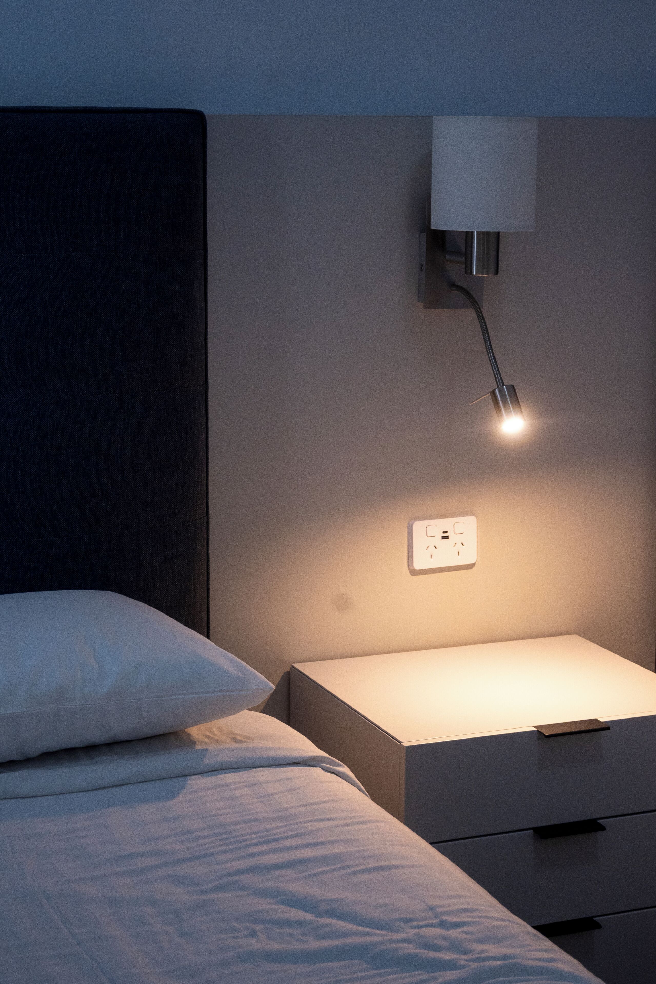 Una cama bien hecha con una almohada, una mesita de noche con una lámpara y un enchufe en la pared.