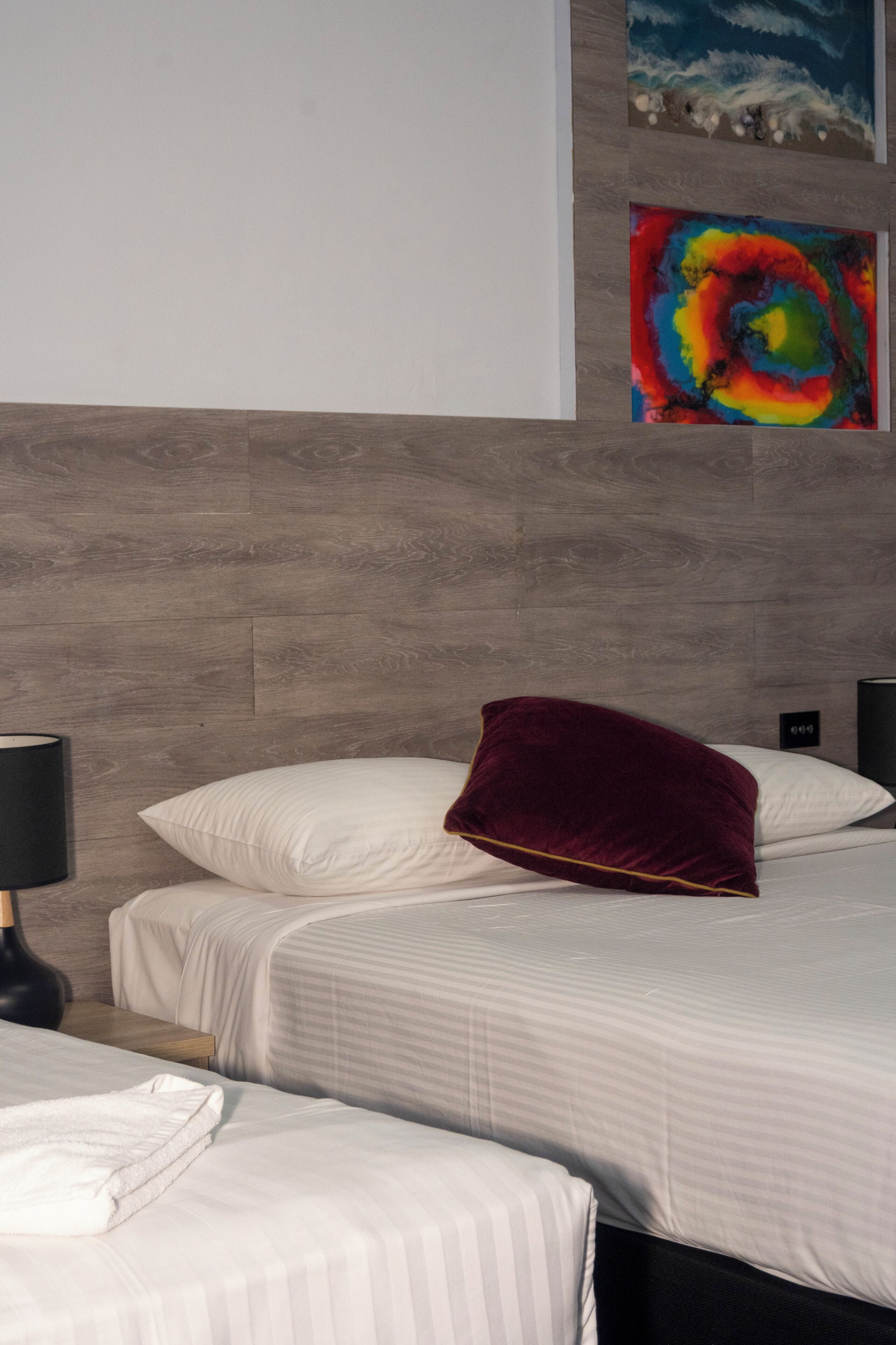 A hotel room with two beds, a wooden headboard, a nightstand with a lamp, and artwork on the wall.