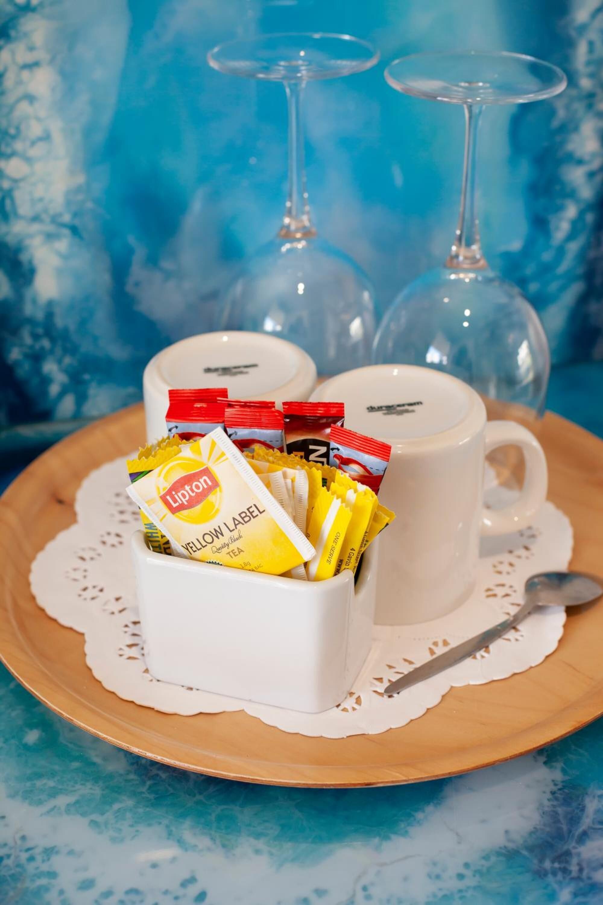 Ein Tablett mit Lipton Teebeuteln und zwei weißen Tassen auf einer Holzschale mit Unterlage, vor blauem Hintergrund.