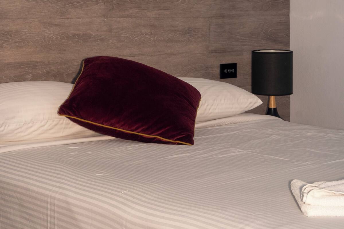 Habitació d'hotel amb un llit, un coixí vermell, una petita taula de nit amb una làmpada i dues obres d'art a la paret.