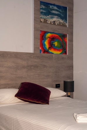 Hotellihuone, jossa on sänky, punainen tyyny, yöpöytä lampun kanssa ja kaksi taideteosta seinällä.
