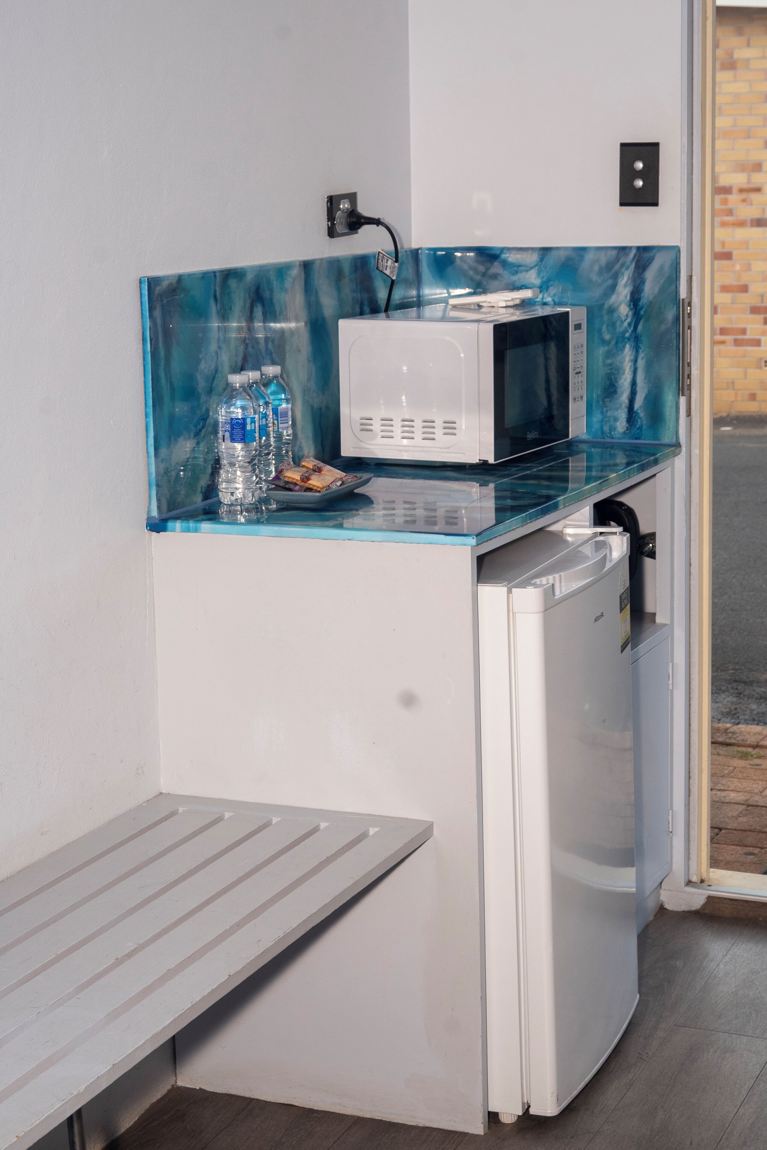 Une petite cuisine équipée d’un four à micro-ondes, d’un petit réfrigérateur et d’un plan de travail en verre sur lequel se trouvent des bouteilles d’eau et une assiette.