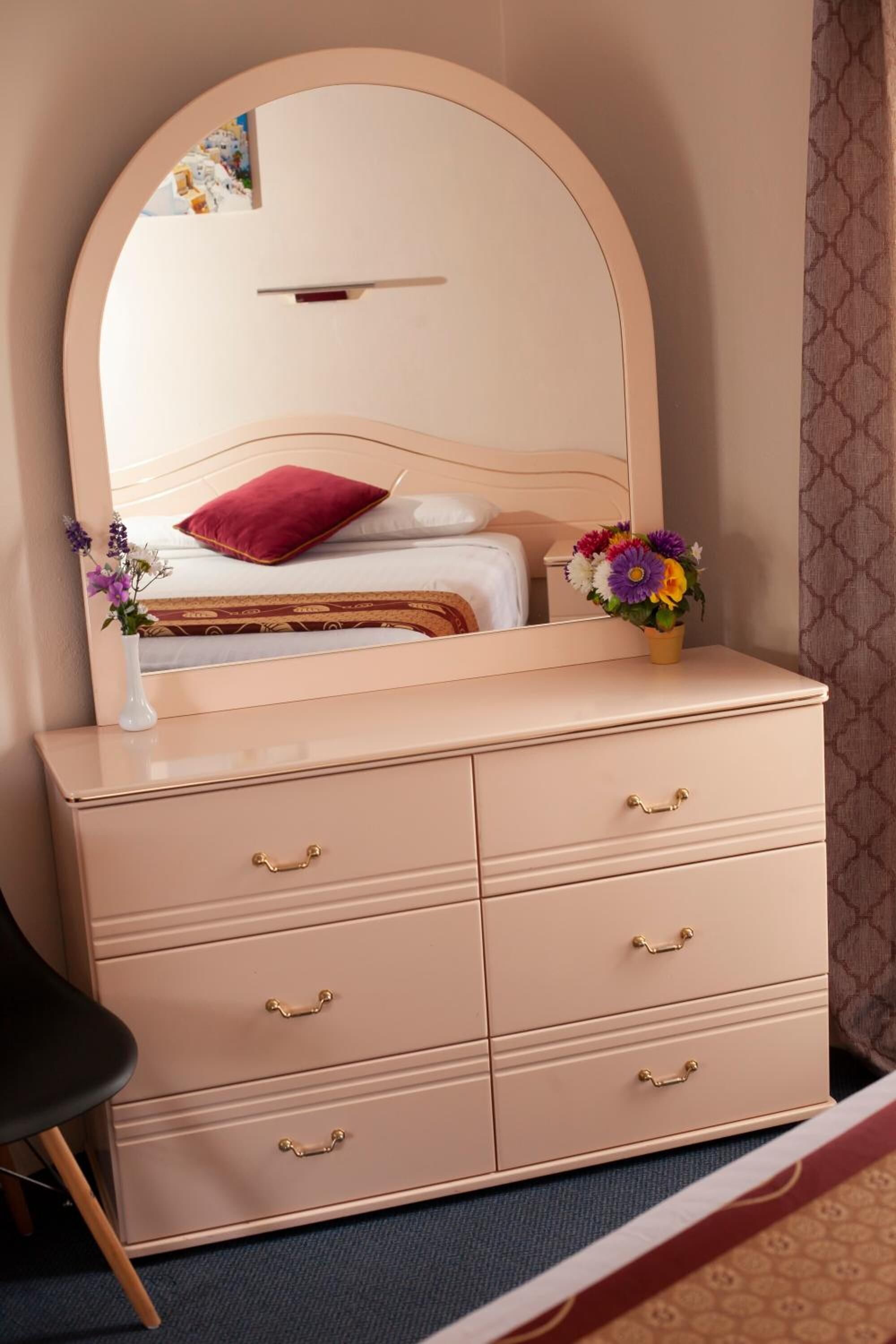 Ein Schlafzimmer mit einem großen Spiegel, einem Bett mit rotem Kopfkissen, einer Kommode mit Schubladen, einer kleinen Vase mit Blumen und einem Sessel.
