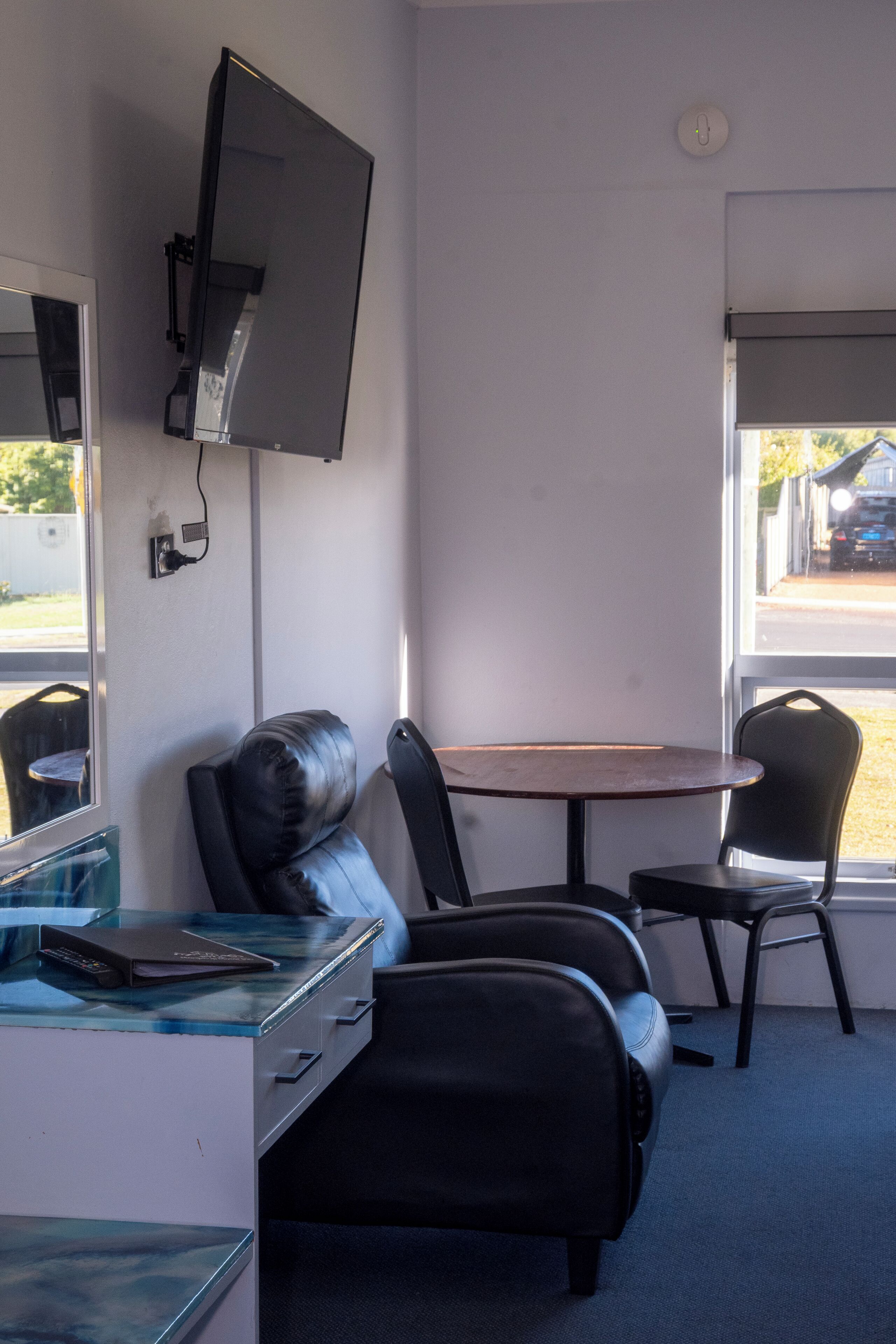 A room with a desk, a chair, a table, a television mounted on the wall, and a window with a view of the outside.