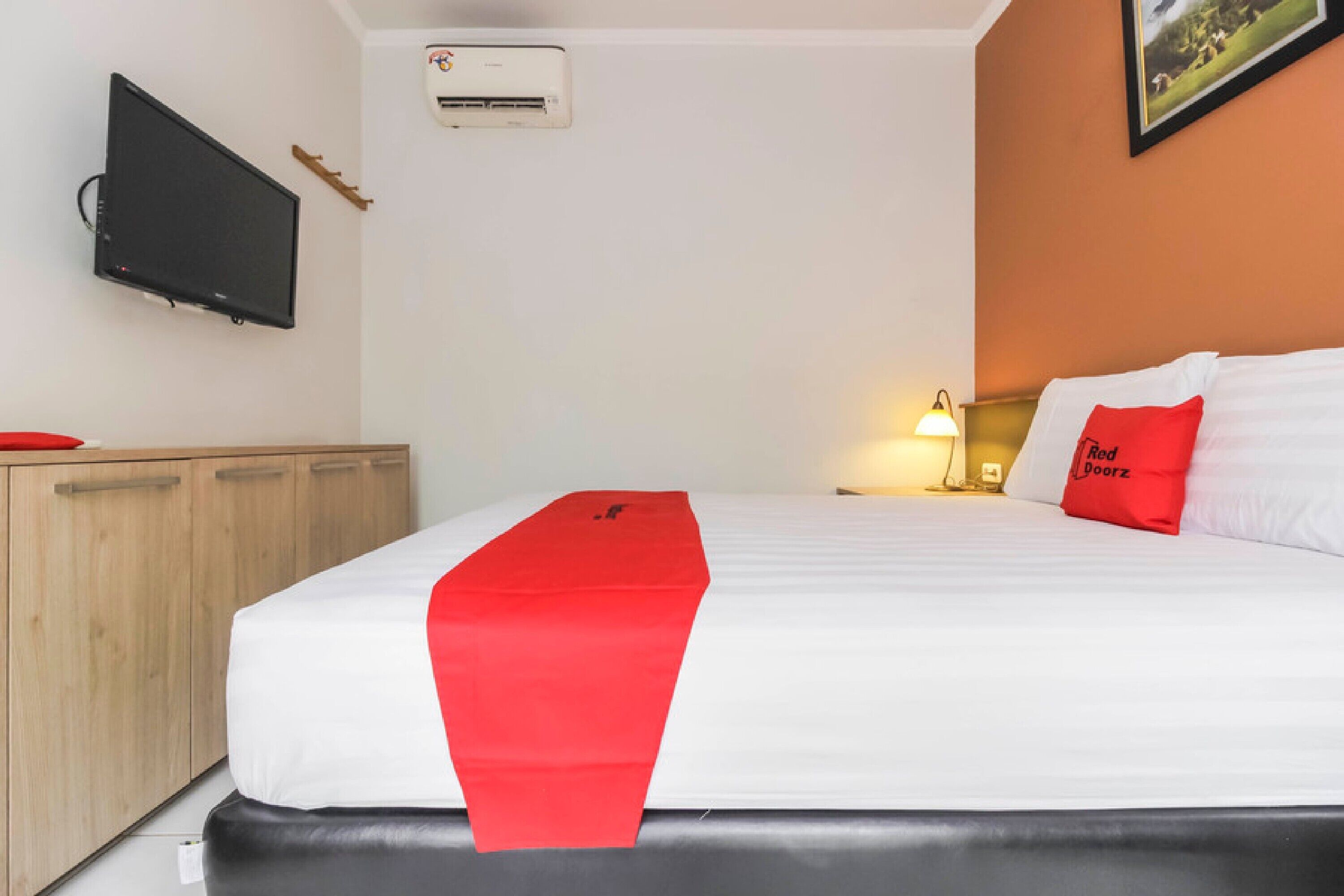 A hotel room with a bed, a TV mounted on the wall, an air conditioning unit, and a lamp on the nightstand.