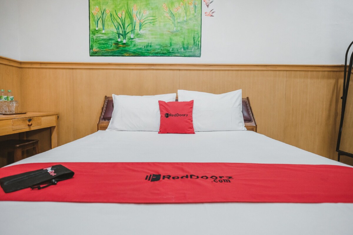 Una cama bien hecha con una manta roja y almohadas, un cabecero de madera, una mesita de noche con una lámpara y un cuadro en la pared.