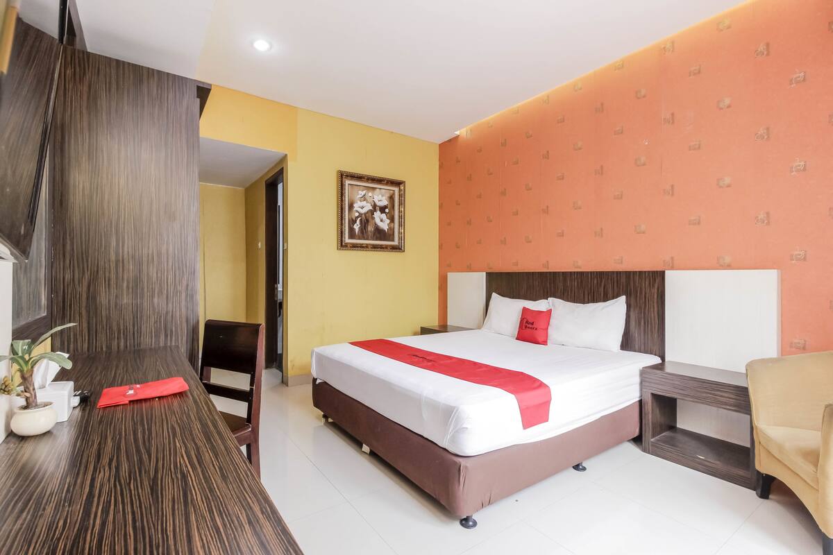 A hotel room with a large bed, a wooden desk, a chair, a small table, and a potted plant.