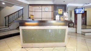 A reception desk with a clock and a wall clock above it, a staircase to the left, and a television on the right. - RedDoorz Plus @ Singosari Raya (Semarang)