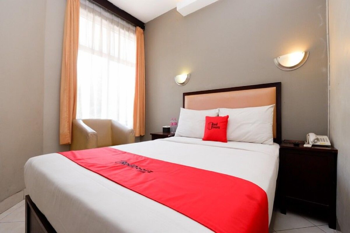 A hotel room with a large bed, a red throw blanket, a nightstand with a phone, and a window with curtains.