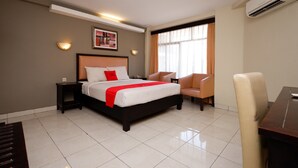 A hotel room with a bed, a desk, a chair, a nightstand, and an air conditioning unit. - RedDoorz Plus @ Singosari Raya (Semarang)