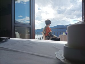 A person seated at a table with a view of a lake and mountains through a window. - AURORA Suite apartment Lake Orta-beach00311600048 (pettenasco)