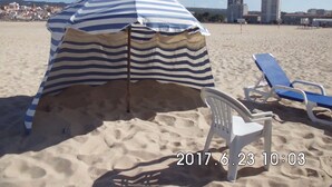 Ein Strandkorb mit gestreiftem Stoff, ein weißer Plastikstuhl und eine blaue Liege an einem sandigen Strand, im Hintergrund sind Gebäude zu sehen.