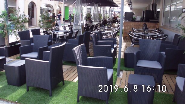 Ein Straßencafé mit Korbmöbeln und Sonnenschirmen, umgeben von viel Grün und einem Gebäude im Hintergrund.