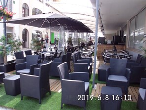 Ein Straßencafé mit Korbmöbeln und Sonnenschirmen, umgeben von viel Grün und einem Gebäude im Hintergrund.