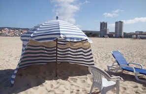 A beach umbrella with striped fabric, a white plastic chair, and a blue lounge chair on a sandy beach with buildings in the background.