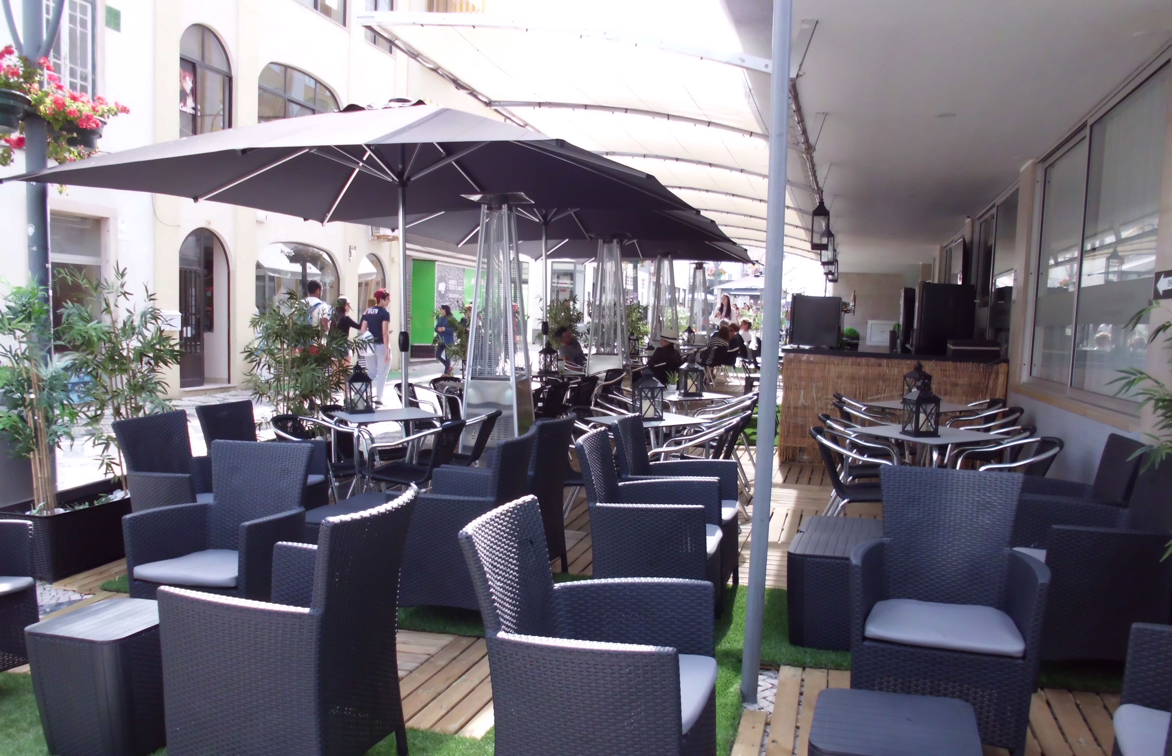 Un café en plein air, avec des meubles en osier et des parasols, entouré de verdure et avec un bâtiment en arrière-plan.