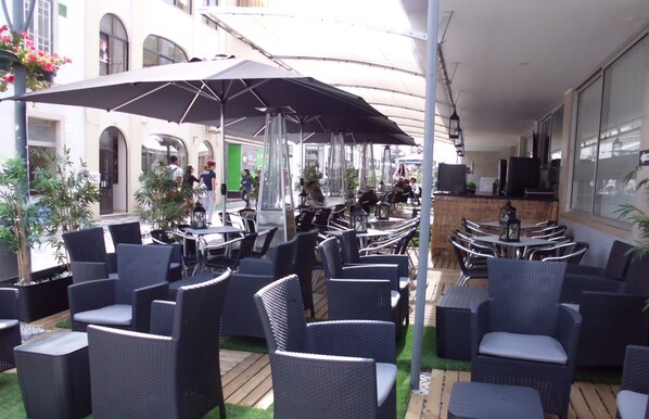 An outdoor cafe with wicker furniture and umbrellas, surrounded by greenery and a building in the background.