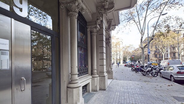 A building with a metal door and large windows, a tiled sidewalk, parked motorcycles, and a car on the street. - Three bedroom apartment with balcony in Ali Bei - You Stylish (Barcelona)