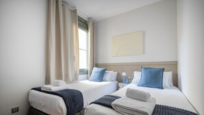 A hotel room with two beds, a window with curtains, a bedside table with a lamp, and a wall-mounted artwork. - Three bedroom apartment with balcony in Ali Bei - You Stylish (Barcelona)