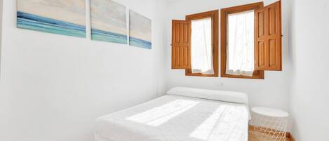 A bedroom with a bed, two windows with wooden shutters, and three wall-mounted paintings.