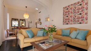 Ein Wohnzimmer mit einem beigefarbenen Sofa, einem Glastisch und einer Vase mit Blumen. An der Wand hängt ein Gemälde und auf dem Beistelltisch steht eine Lampe.