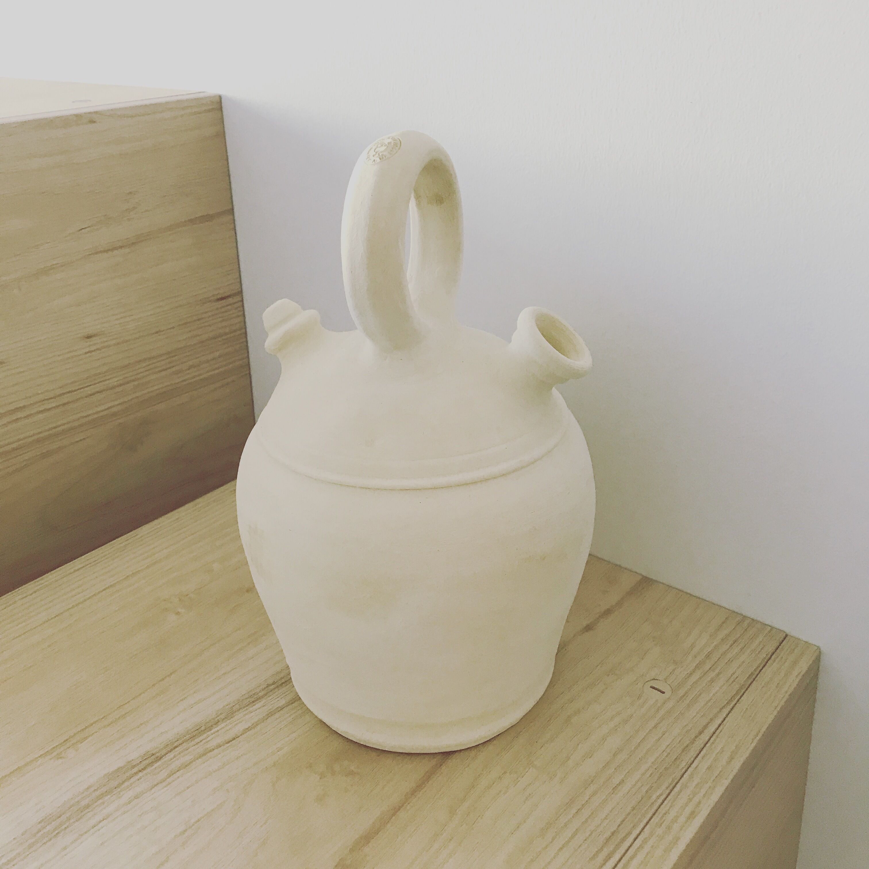 Una jarra de cerámica blanca con asa y pico, sobre una superficie de madera frente a una pared lisa.