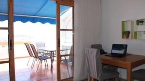 A room with a wooden table, a laptop, and a view of an outdoor patio with chairs and a table.
