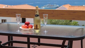 A table with a bottle of champagne, two glasses, and a bowl of fruit on a balcony overlooking the sea.