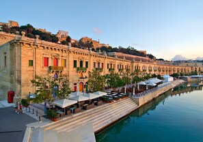 A waterfront hotel with outdoor seating along a canal, stone buildings with balconies, and a view of the cityscape. - Seafront Lux Fort Cambridge wt Pool (Sliema)