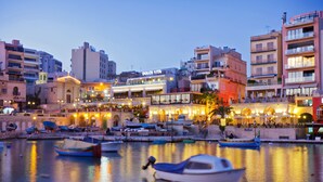 A waterfront area with boats docked, buildings with shops, and a prominent Dolce Vita sign. - Seafront Lux Fort Cambridge wt Pool (Sliema)