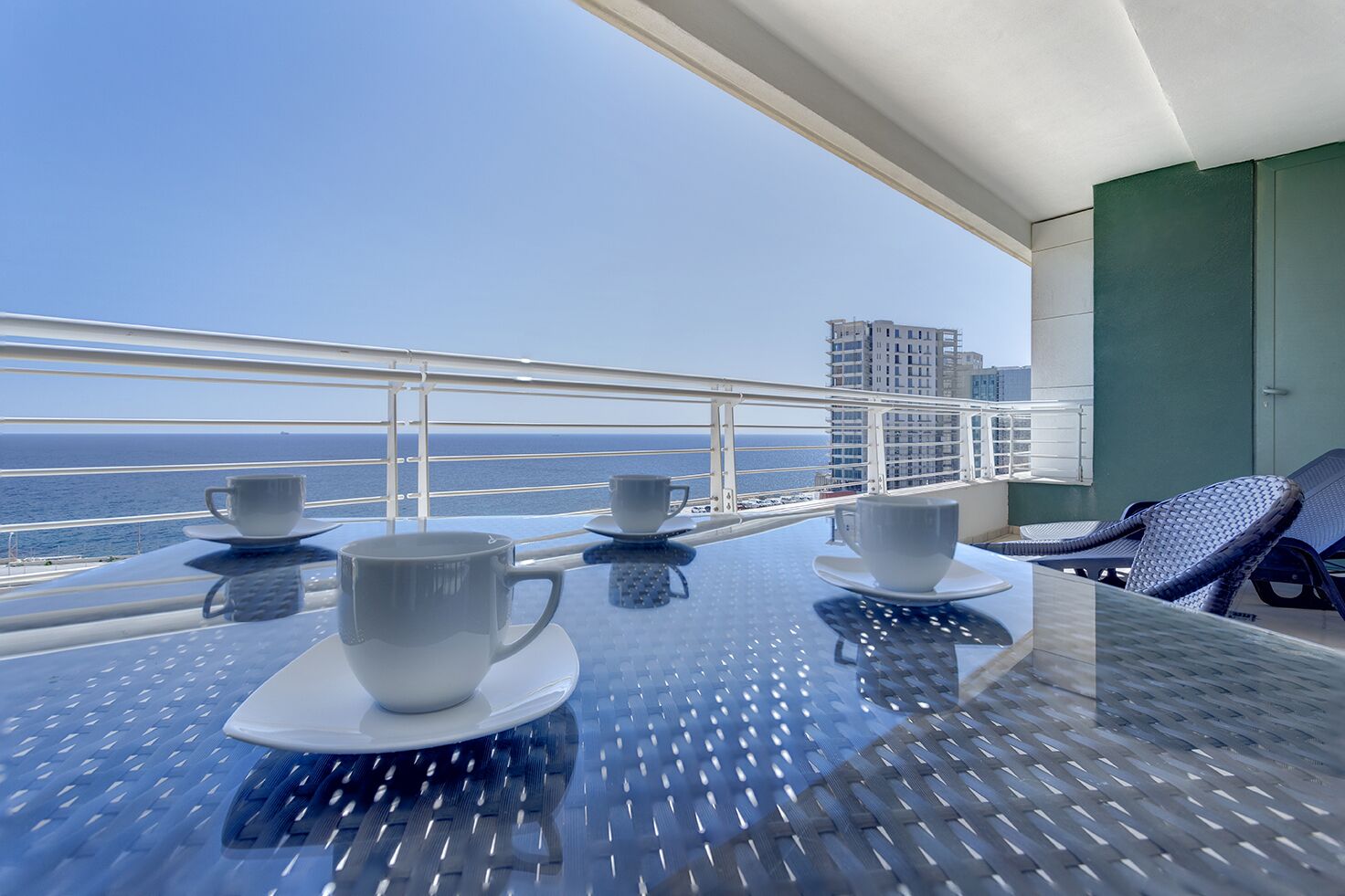 A balcony with a table set for two, two cups, and a view of the ocean and buildings.