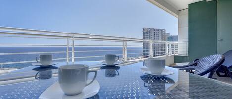 A balcony with a table set for two, two cups, and a view of the ocean and buildings.