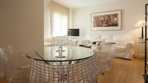 Un salon moderne avec une table à manger au plateau en verre, entourée de chaises au design épuré, d’un canapé blanc et d’un téléviseur à écran plat.