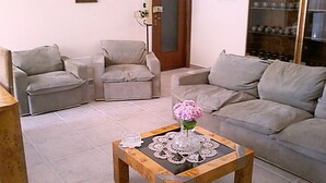 Una sala de estar con un sofá gris, dos sillones, una mesa de centro de madera con un florero, y piso de baldosas.