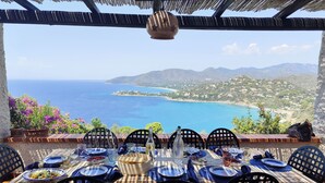 Un espace de restauration en plein air avec une table dressée pour quatre personnes, offrant une vue sur la côte et les montagnes au loin.