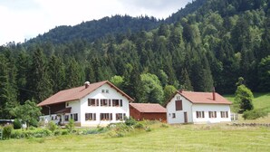 A white building with a red roof and a small attached structure, surrounded by a grassy field and a forested hill.