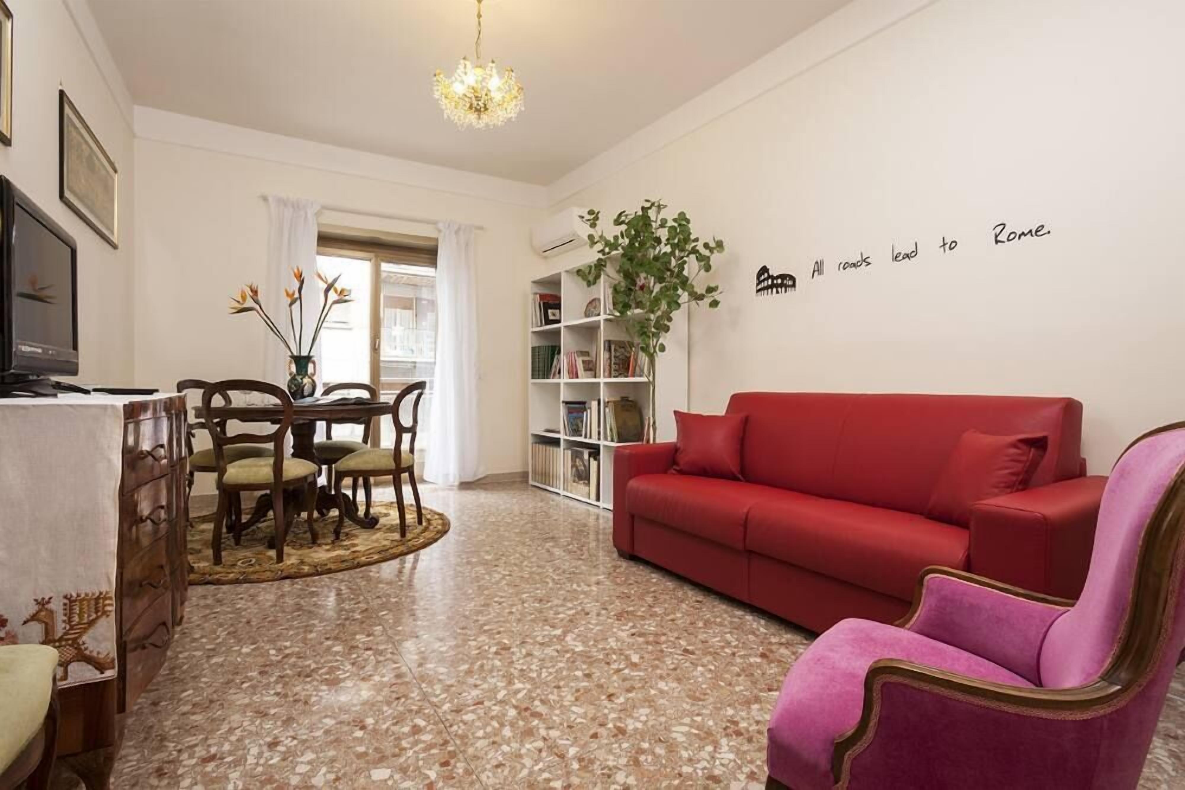 Ein Wohnzimmer mit einem roten Sofa, einem Esstisch mit Stühlen, einer Chandelüster und einem Bücherregal.