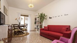 Uma sala de estar com um sofá vermelho, uma mesa de jantar com cadeiras, um candelabro, uma estante e uma parede com uma citação.