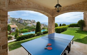 Ein Tischtennis-Tisch mit zwei Schlägern in einem Steingewölbe mit Blick auf einen Pool und Gebäude am Hang.