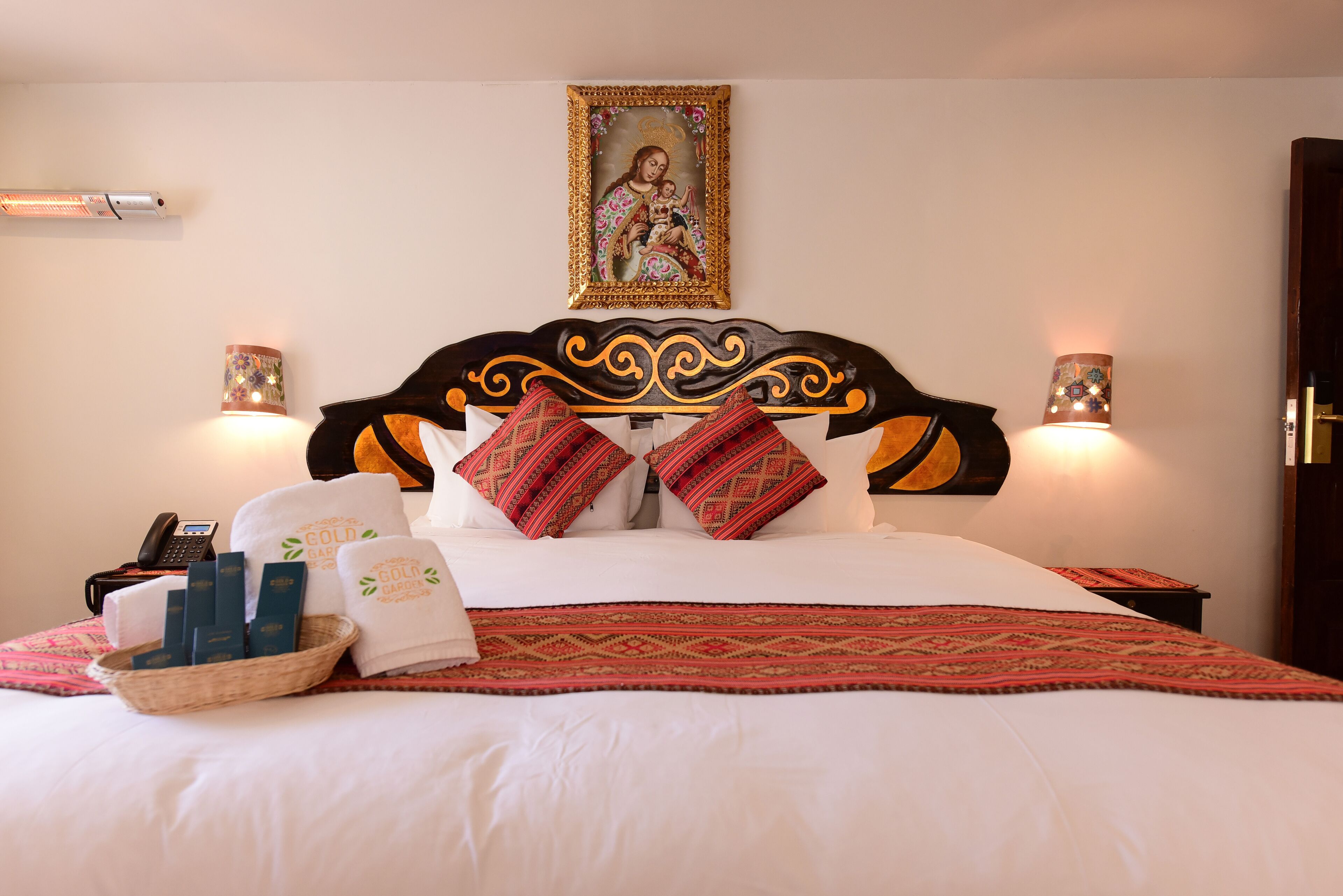 A neatly made bed with a patterned bedspread, two decorative pillows, a framed religious painting above the bed, and two wall-mounted lamps.