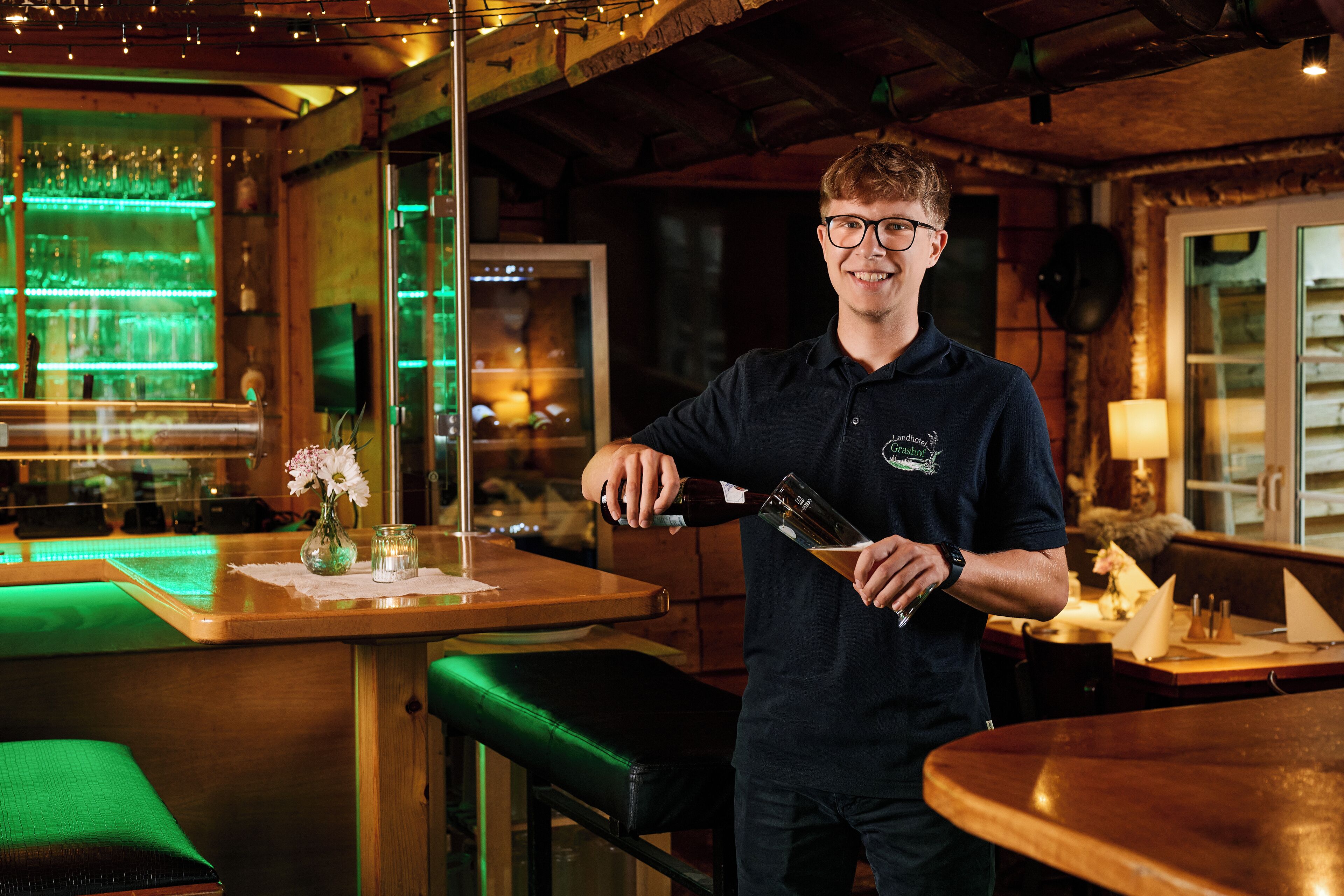 A person pouring a drink at a bar with a green-lit bar counter and wooden interior.