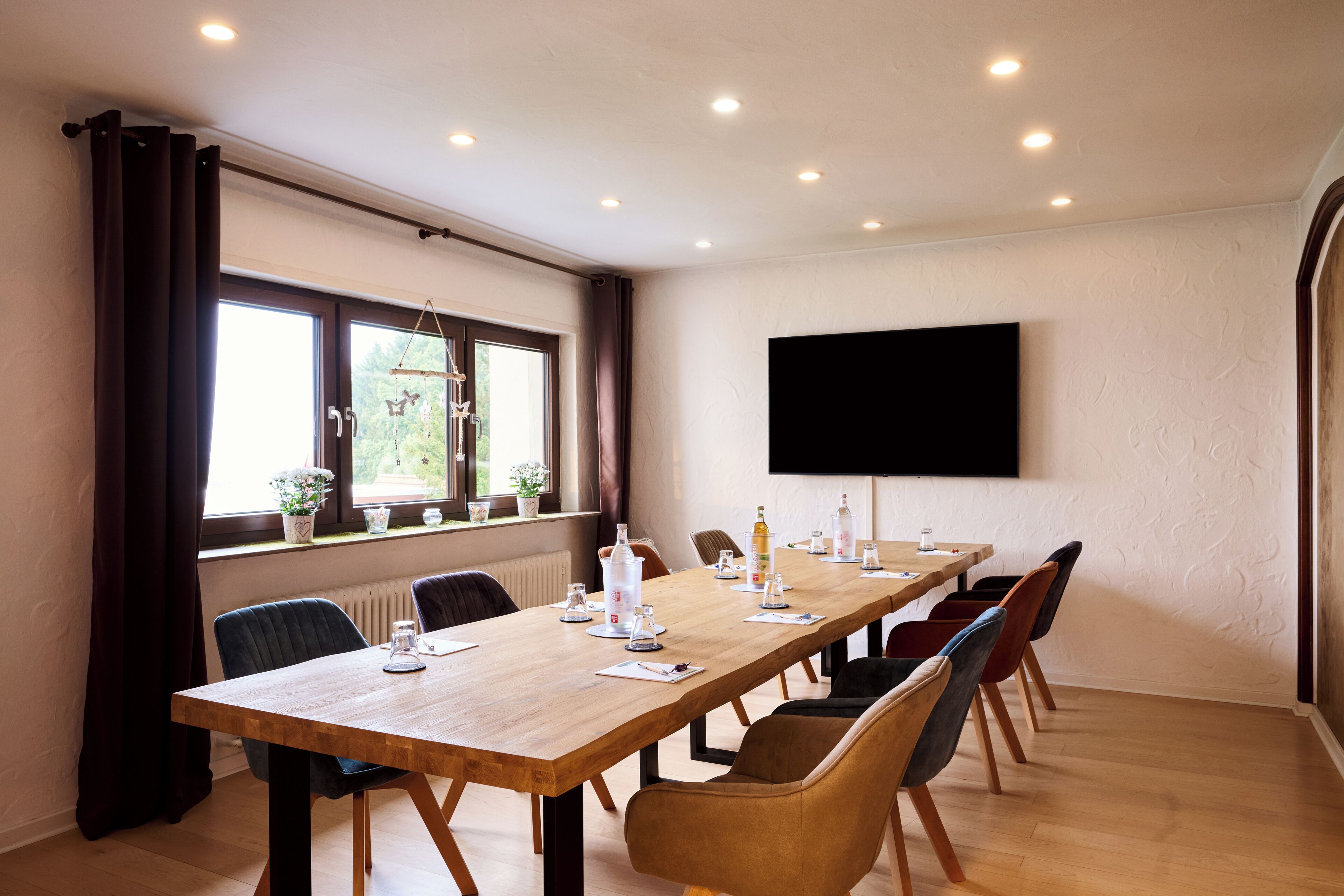 A conference room with a long table, chairs, a flat-screen TV, and windows with curtains.