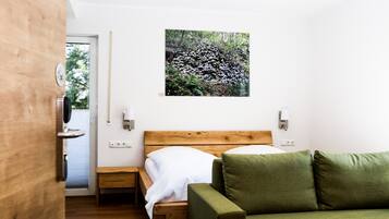 A bedroom with a wooden headboard bed, a green sofa, a wall-mounted picture of a forest, and a small bedside table with a lamp.