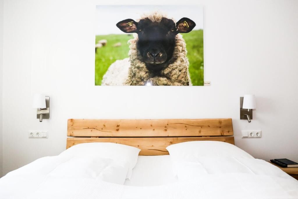 Een hotelkamer met een houten hoofdeinde, een bed met wit beddengoed, een schilderij van een schaap aan de muur, en twee nachtlampjes.