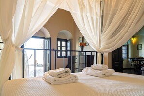 A bedroom with a bed, white curtains, a view of the outside, and a vase with flowers.