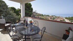 A balcony with a round table and chairs, potted plants, and a view of the sea and town.