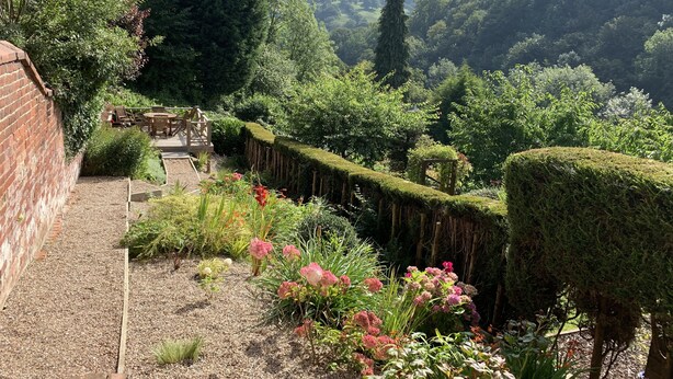 Un jardí amb mur de maó, terrassa de fusta i diferents plantes i flors.