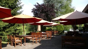 An outdoor patio area with wooden tables and chairs, red and beige umbrellas, and a brick floor. - Flörsbacher Hof (Floersbachtal)